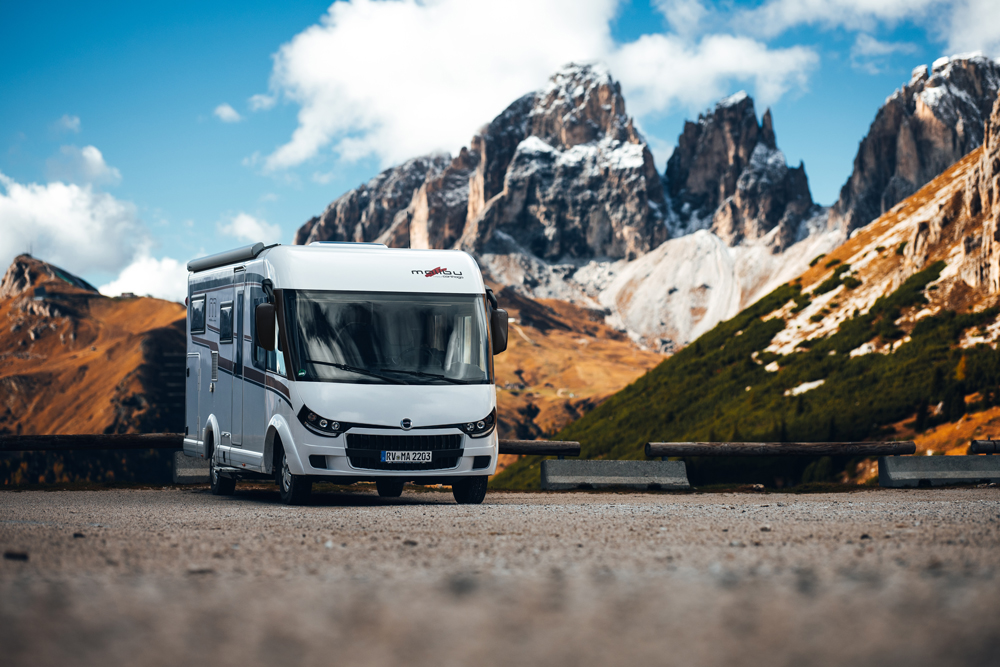 Vollintegriertes Malibu Wohnmobil auf Basis eines Fiat-Fahrgestells, geparkt vor einer ländlichen Kulisse mit Bäumen und Bergen im Hintergrund. Das Fahrzeug zeigt seine stilvolle Frontpartie und aerodynamisches Design.