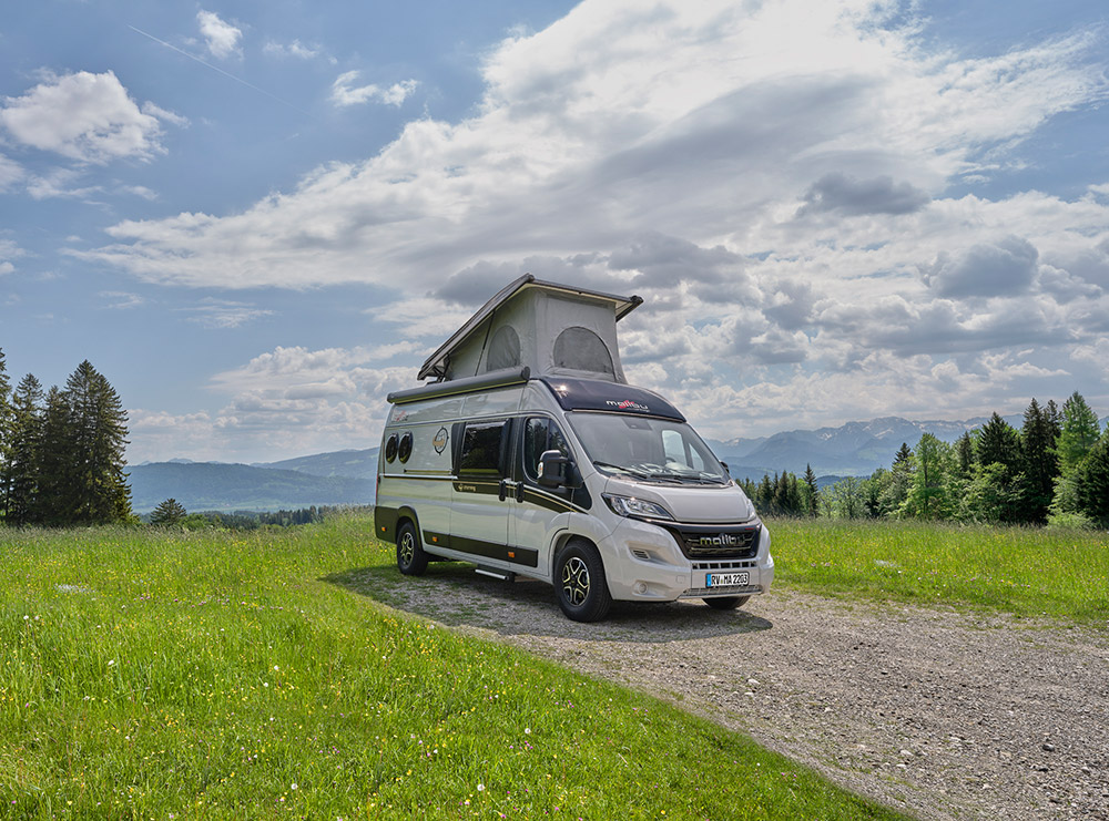 Der Malibu Van mit aufgeklapptem Schlafdach steht auf einer grünen Wiese, umgeben von sanften Hügeln und einem malerischen Bergpanorama unter einem leicht bewölkten Himmel.