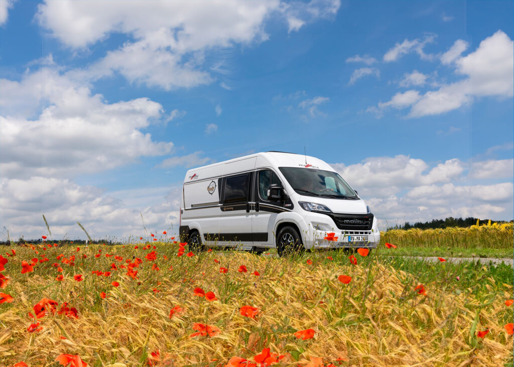 Weißer Malibu Van fährt eine Straße entlang, schönes wetter, Felder und Blumen Blühen.