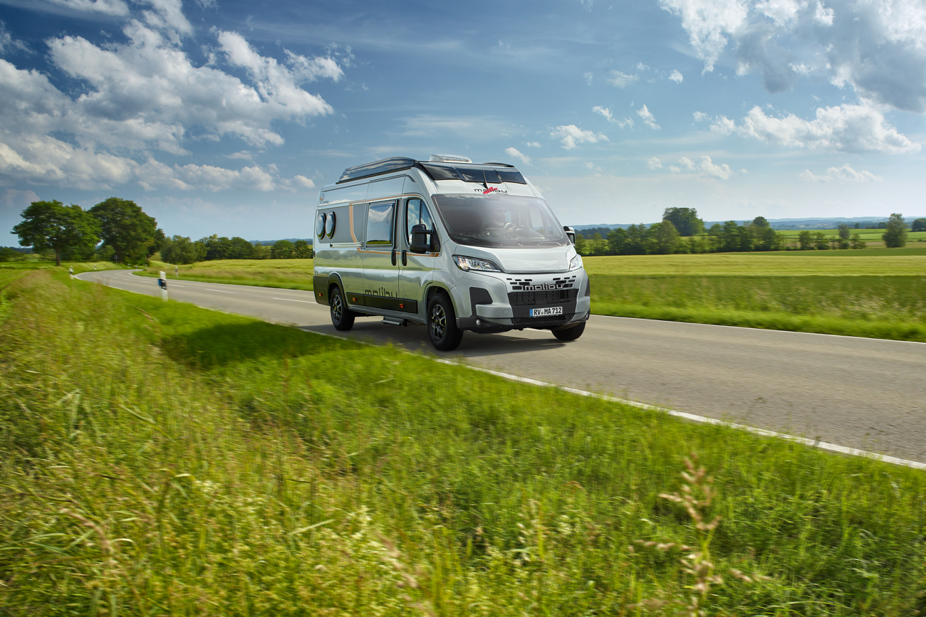 Malibu Van compact 600 LE auf einer Landstraße. Der Van fährt auf einer Straße, umgeben von Wolken und hohen Gräsern, mit Feldern und Bäumen im Hintergrund.