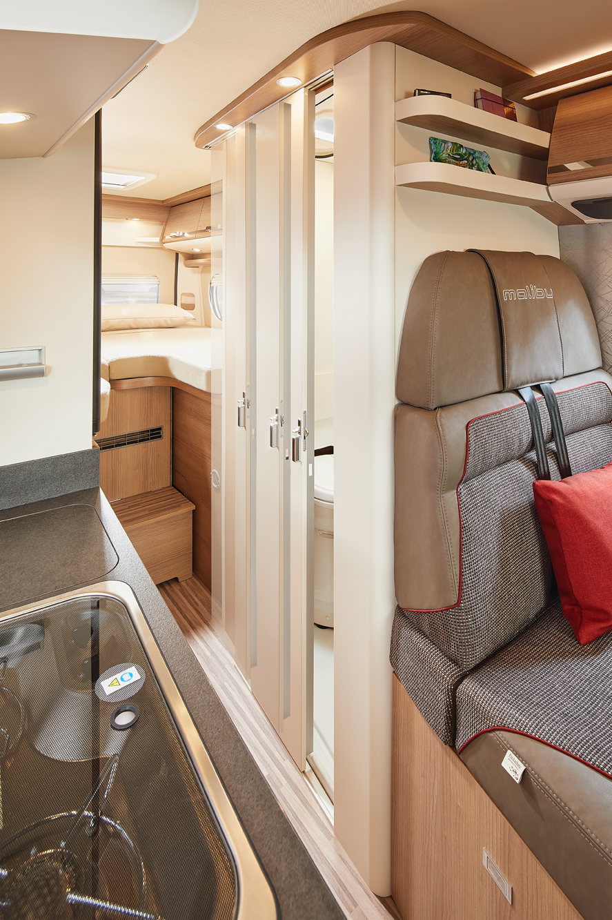 stilvolle und funktionale Interieur eines Malibu Vans, mit hochwertig gepolsterten Sitzen, einer Küchenzeile im Vordergrund und einem Schlafbereich im Hintergrund. Klappbare Türen trennen den Wohnbereich von einem kompakten Badezimmer, während warme Holztöne und dezente Beleuchtung eine gemütliche Atmosphäre schaffen.