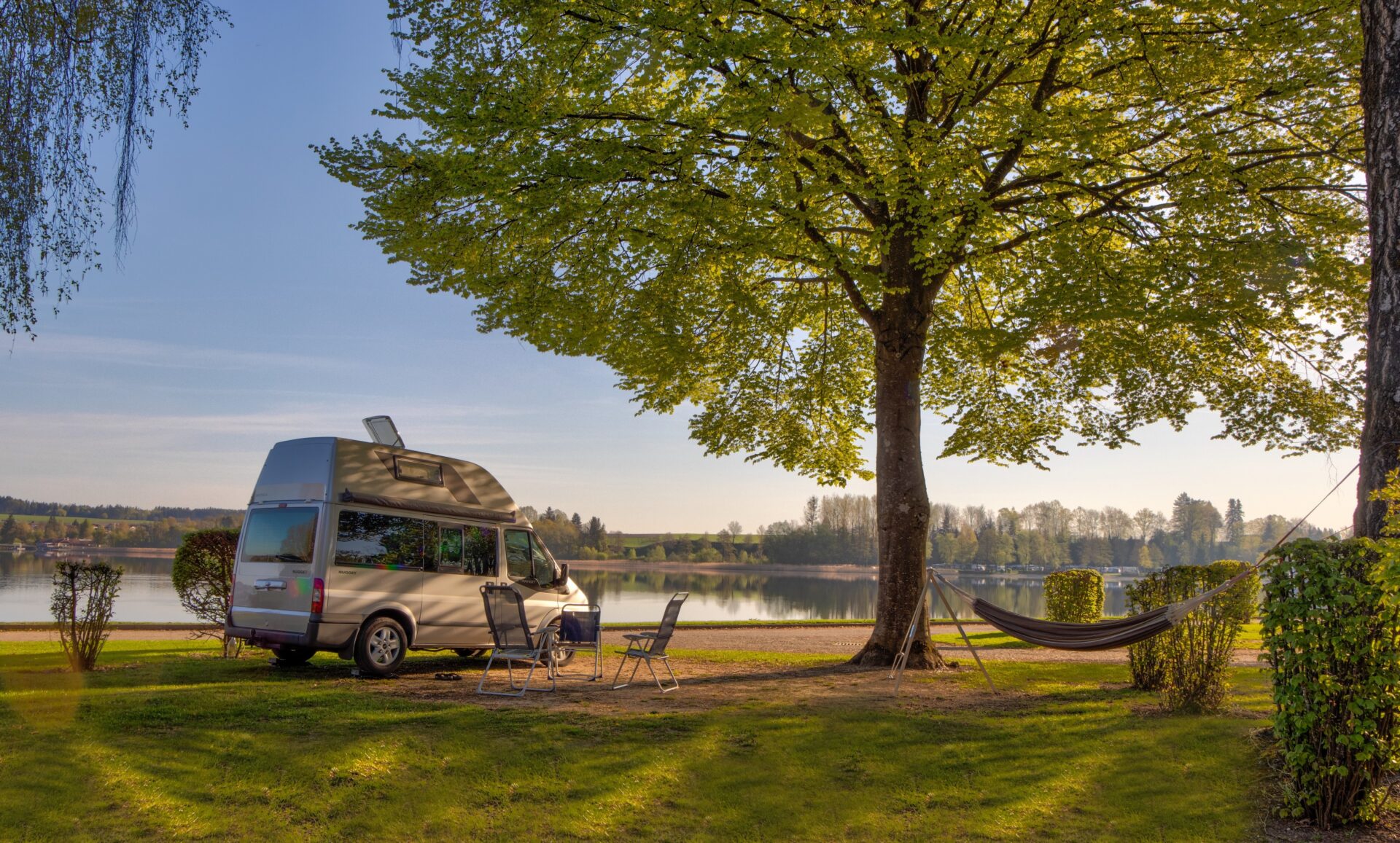 Ein Camper Van unter einem großen grünen Baum bei Sonnenschein mit zwei Liegestühlen direkt am See.