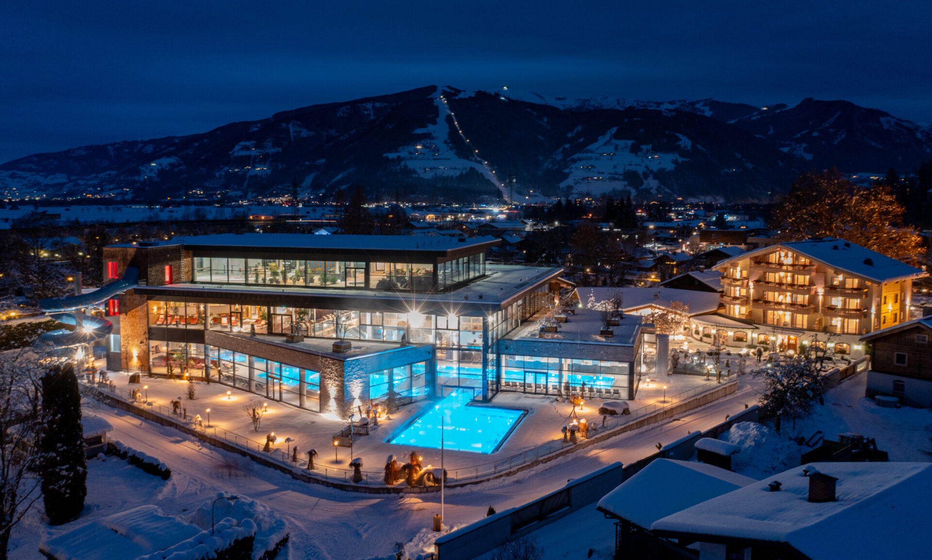 Nachtaufnahme von einem beleuchteten Hotel mit Pool umgeben von schneebedeckten Bergen