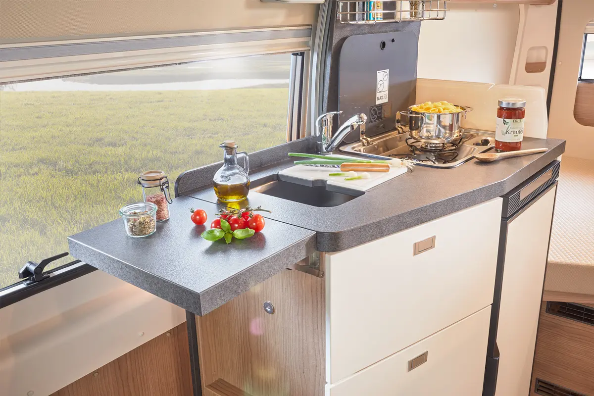 Küchenarbeitsfläche mit frischem Gemüse, Ölflasche und Kochutensilien. Fenster mit Blick auf grüne Landschaft.