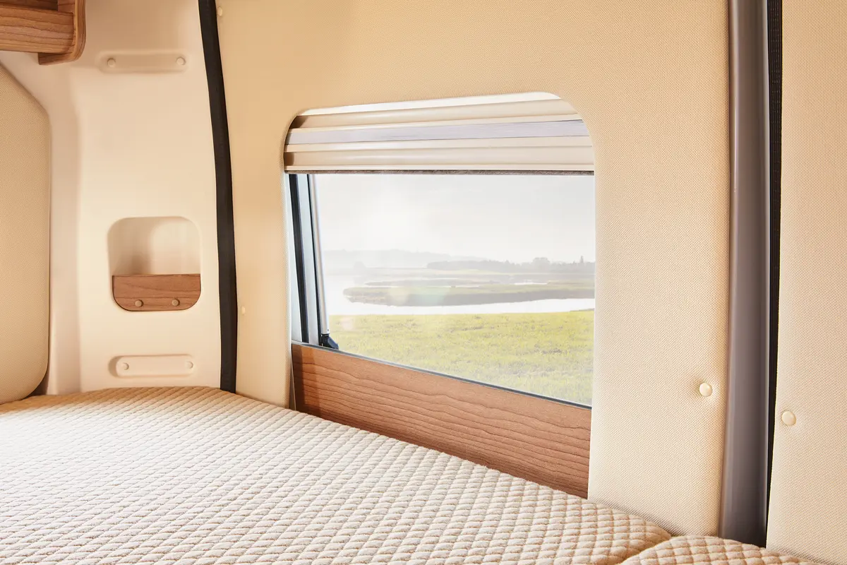 Innenansicht eines Vans mit einem gemütlichen Bett, das unter einem Fenster mit Blick auf eine grüne Landschaft platziert ist. Die weichen Beigetöne der Einrichtung und die Holzdetails schaffen eine warme Atmosphäre.