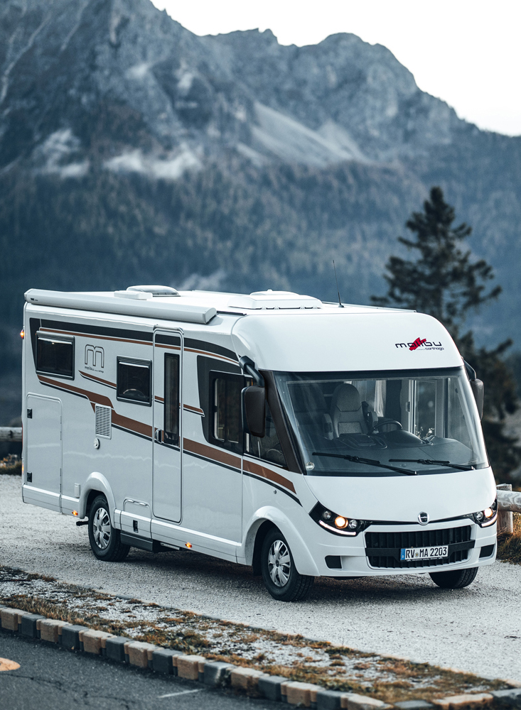 Vollintegriertes Malibu Wohnmobil in einer modernen Außenansicht, geparkt vor einer malerischen Kulisse mit Bergen im Hintergrund. Das Design des Wohnmobils betont seine aerodynamische Front und die großzügigen Fensterflächen.