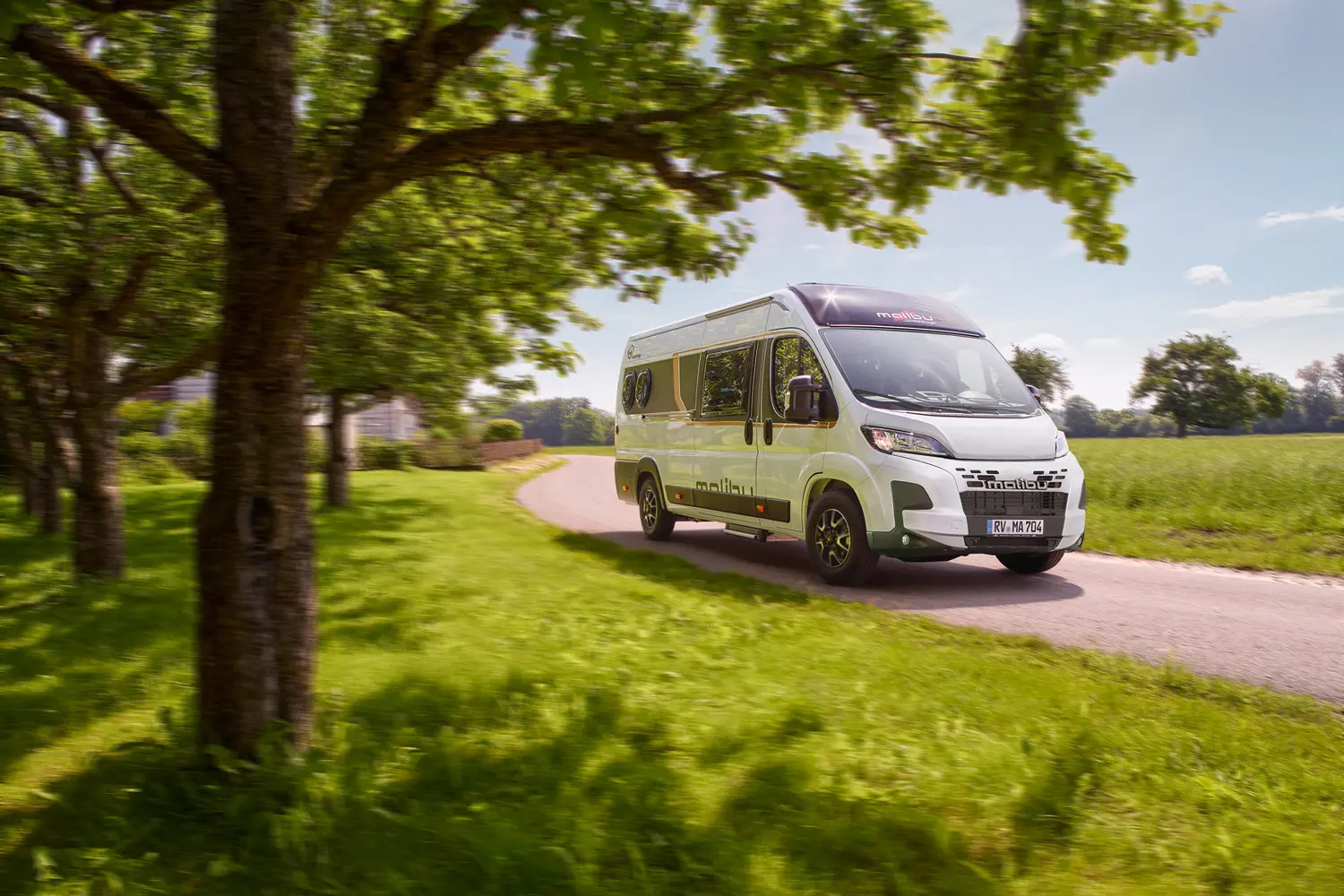 Malibu Van First Class Two Rooms Modell 2025 während der Fahrt, frontal und seitlich aufgenommen, auf einer Straße in einer ländlichen Gegend mit blühenden Feldern und Hügeln im Hintergrund. Das Fahrzeug zeigt seine moderne, dynamische Optik und das GT Skyview-Dachfenster.