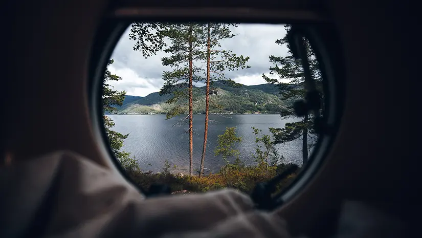 Blick durch ein rundes Fenster auf eine idyllische Naturlandschaft mit einem See und Bergen im Hintergrund.