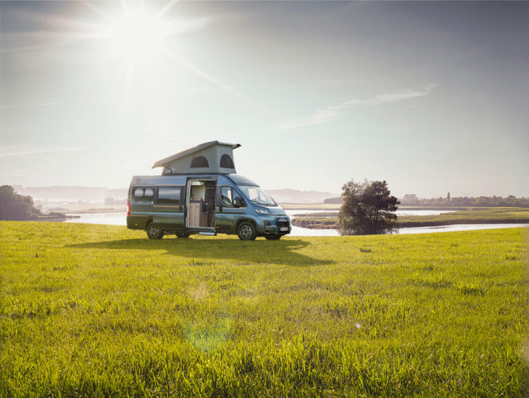 Seitenansicht eines Malibu Campervans mit aufgestelltem Dach auf einer grünen Wiese mit See im Hintergrund.