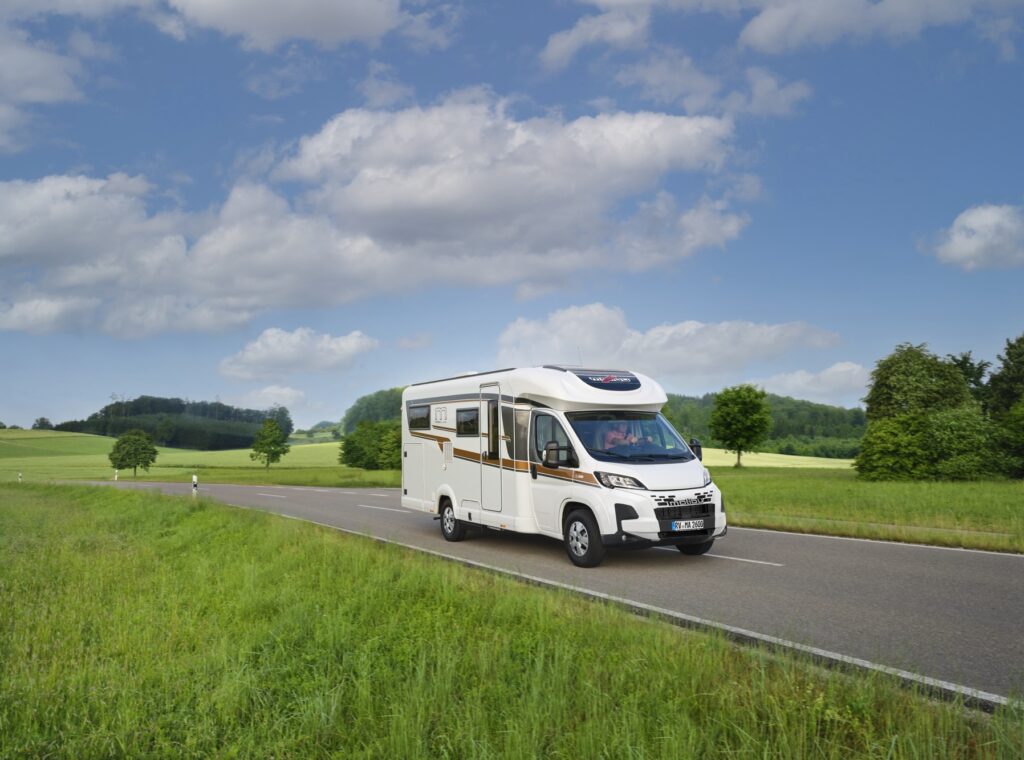 Malibu Van First Class Two Rooms Modell 2025 während der Fahrt, frontal und seitlich aufgenommen, auf einer Straße in einer ländlichen Gegend mit blühenden Feldern und Hügeln im Hintergrund. Das Fahrzeug zeigt seine moderne, dynamische Optik und das GT Skyview-Dachfenster.