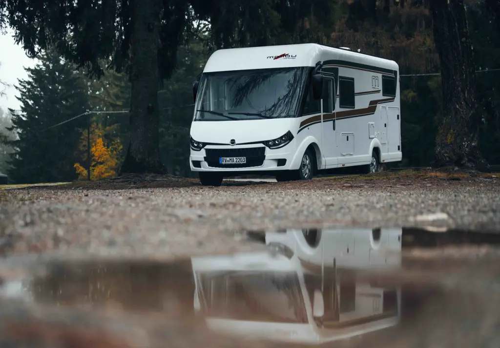 Malibu Reisemobil auf einem Schotterplatz, im Hintergrund Wald, im Vordergrund eine Pfütze